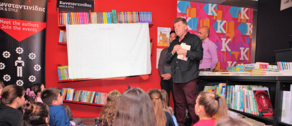 mr papaioanu talks to the students of the 4th grade of mandoulides schools, who sit on the floor with their backs turned from the camera, listening to him