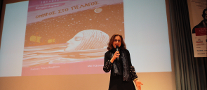 Η κ. Δημότα στη σκηνή του θεάτρου των Εκπαιδευτηρίων, μιλάει κρατώντας ένα μικρόφωνο και πίσω της φαίνεται μια εικόνα με το εξώφυλλο του βιβλίου της