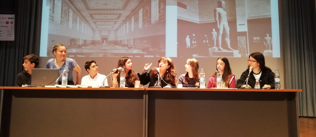 Η συγγραφέας Αγγελική Δαρλάση στην σκηνή του κέντρου εκδηλώσεων των εκπαιδευτηρίων με ένα γραφείο με 7 μαθητές, μιλάει με το κοινό για θέματα που τους απασχολούν