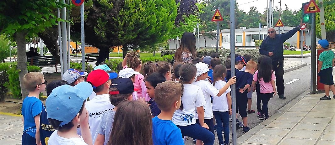 Τα Νήπια των Εκπαιδευτηρίων, με αφορμή τη θεματική ενότητα «Μαθαίνω να κυκλοφορώ με ασφάλεια», επισκέφθηκαν το Πάρκο Κυκλοφοριακής Αγωγής Καλαμαριάς στέκονται σε μια γραμμή όπου ένας κύριος τους δείχνει ένα φανάρι